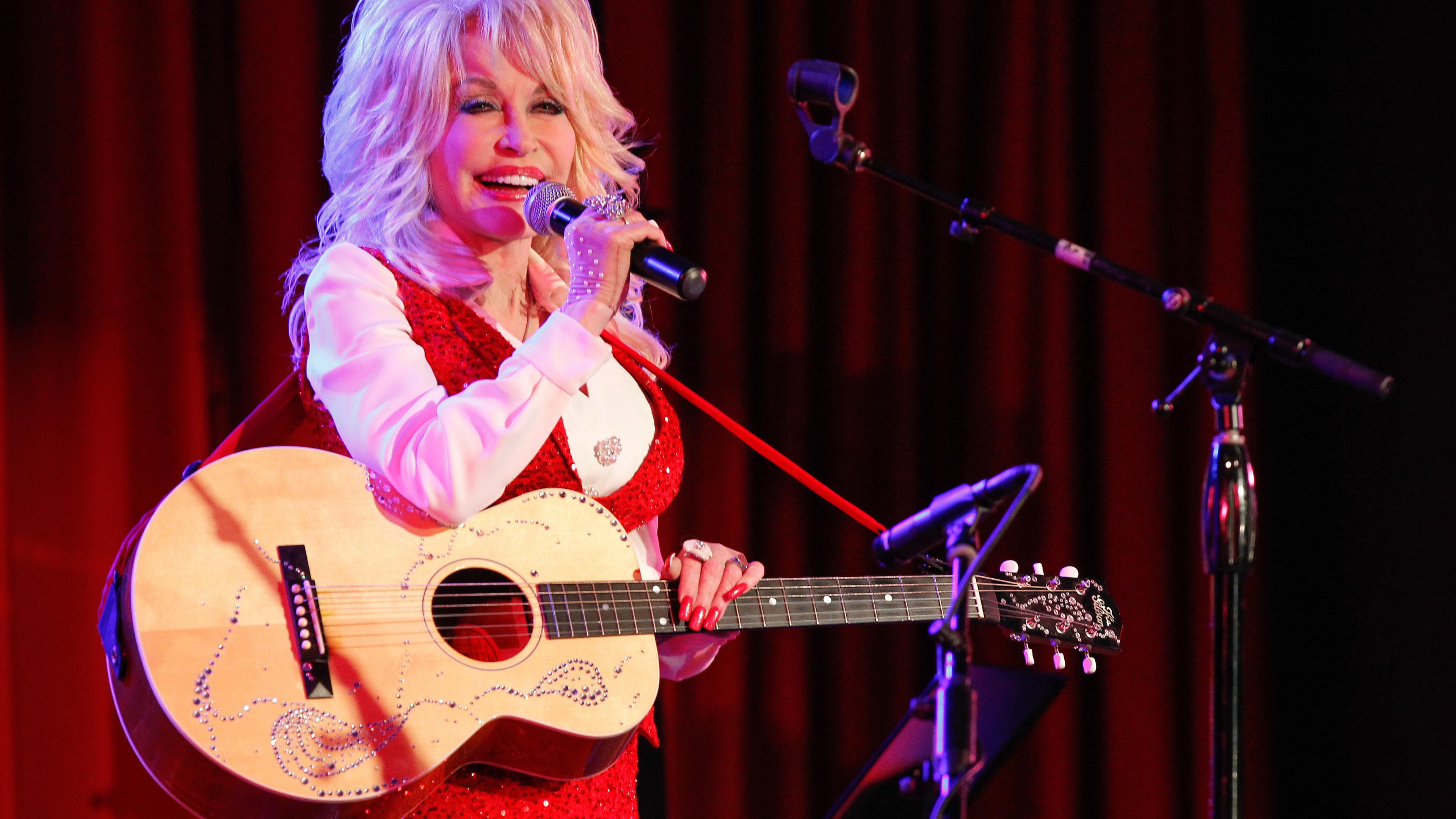 Dolly Parton hält in der linken Hand eine Gitarre. Sie steht vor einem roten Vorhang und singt in ein Mikrofon.