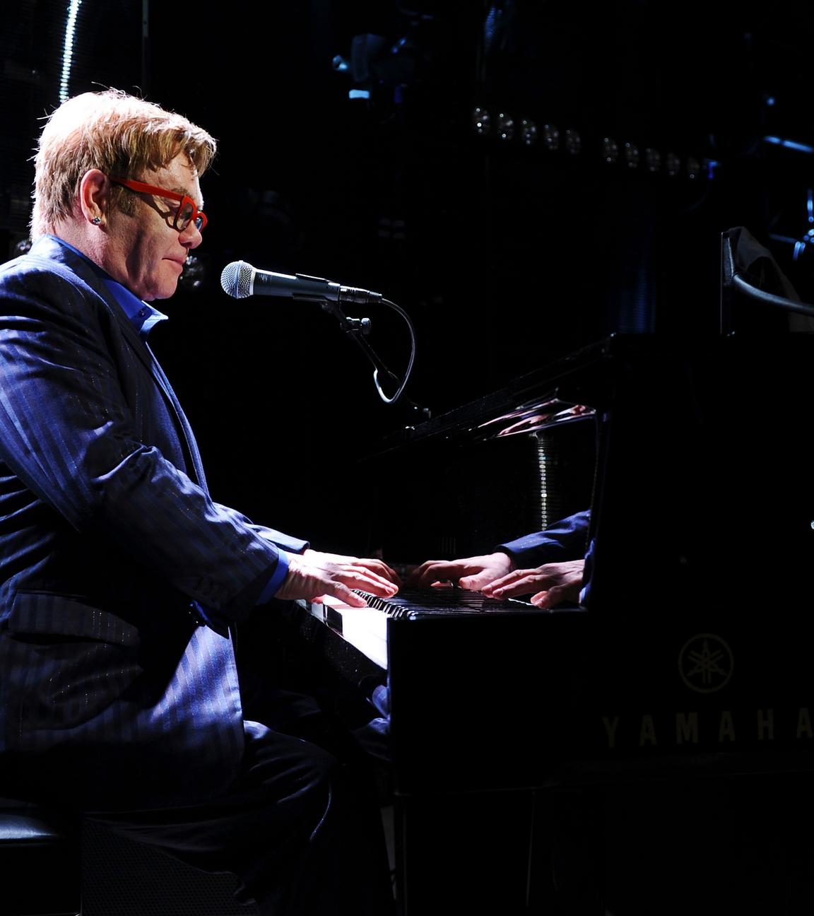 Elton John trägt eine rote Sonnenbrille und ein gestreiftes blaues Sakko. Er spielt Klavier.