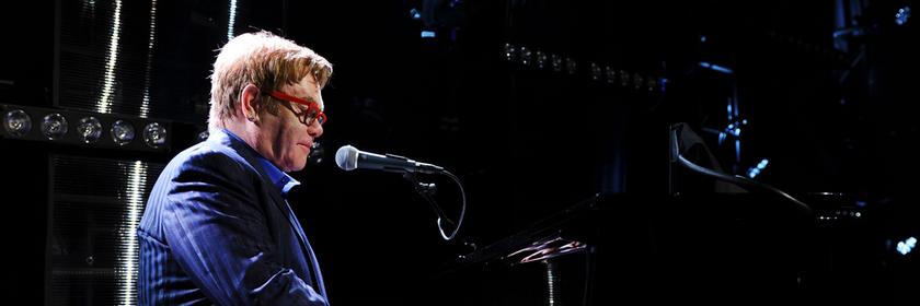 Elton John trägt eine rote Sonnenbrille und ein gestreiftes blaues Sakko. Er spielt Klavier.