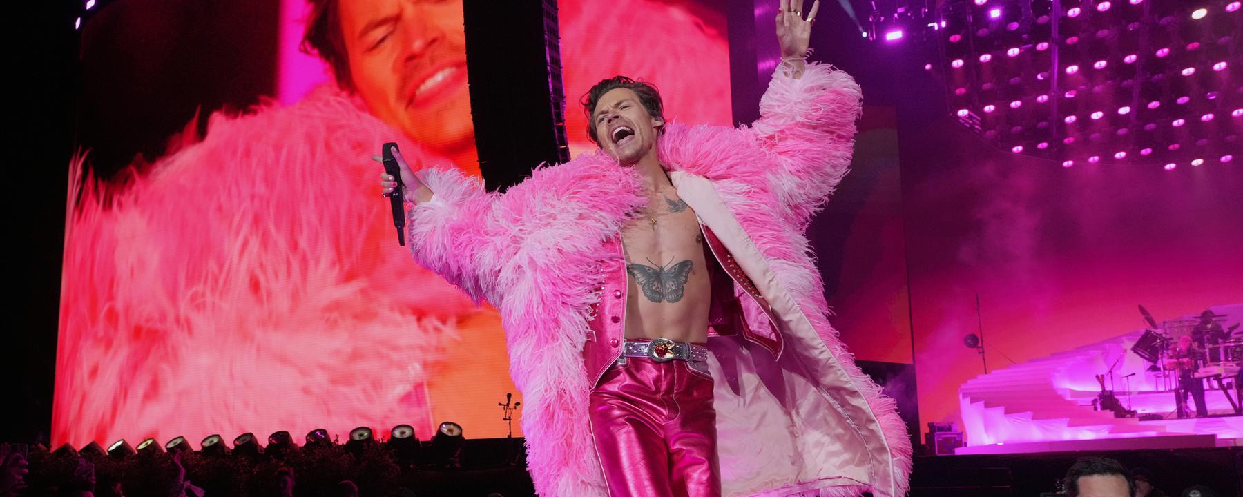 Harry Styles in pinker Metallic-Hose und pinkem Plüschmantel auf einer Bühne. Im Hintergrund Scheinwerfer und Übertragungsleinwand.