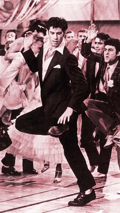 Ein Mann im Anzug, John Travolta, mit offenem Kragen tanzt inmitten einer jubelnden Menge.