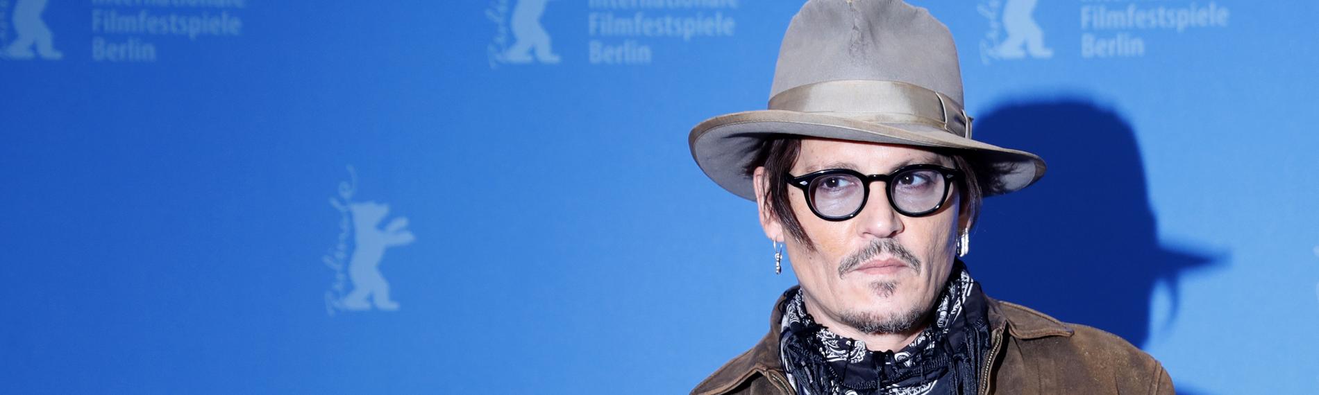 Johnny Depp steht, mit Hut und schwarzer Brille, vor einer blauen Wand.
