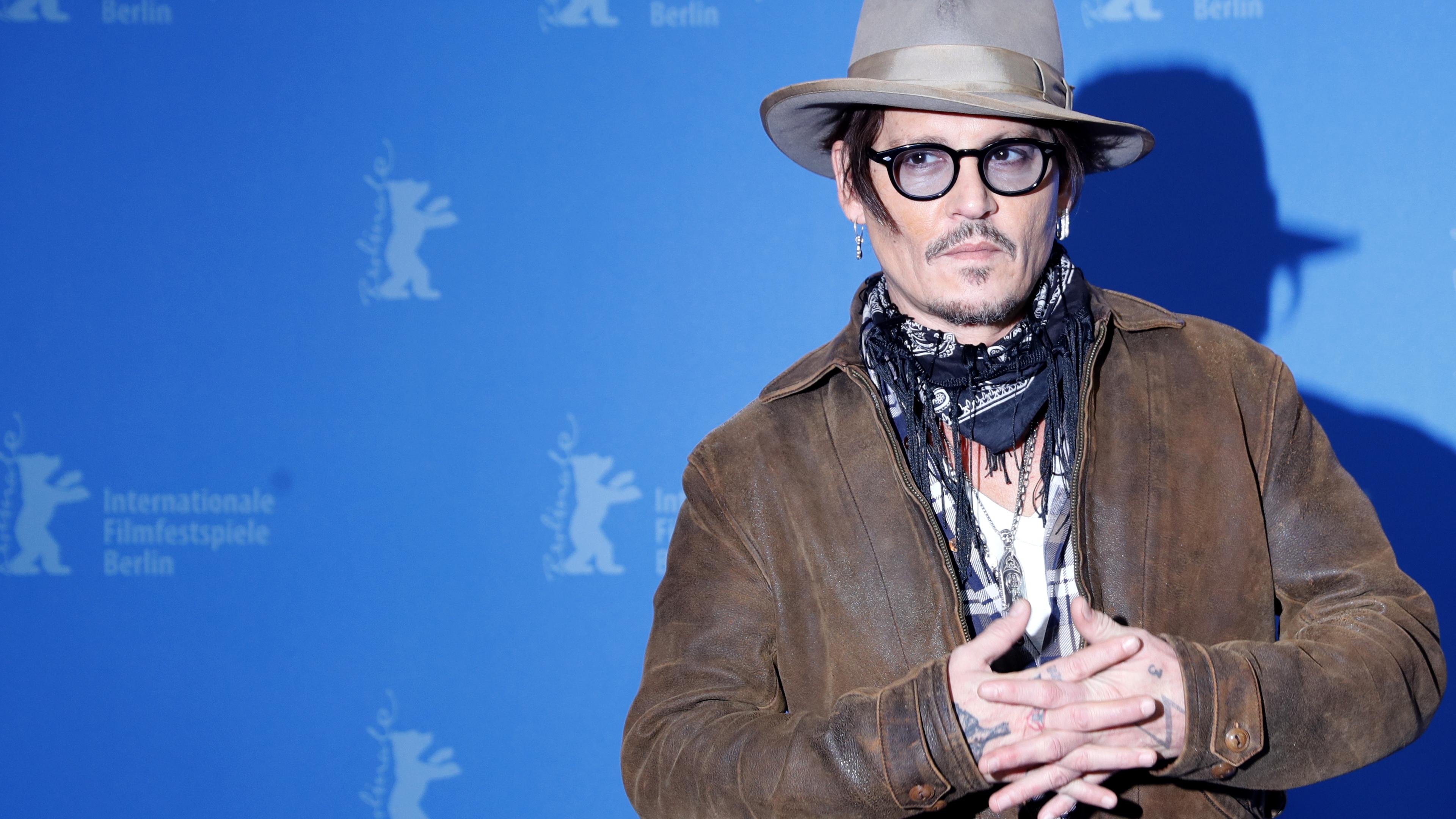 Johnny Depp steht, mit Hut und schwarzer Brille, vor einer blauen Wand.