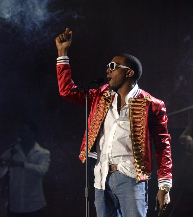 Rapper Kanye West steht auf der Bühne vor einem Mikrofon und singt. Die Bühne ist dunkel, an der Seite leuchtet grelles Scheinwerferlicht.