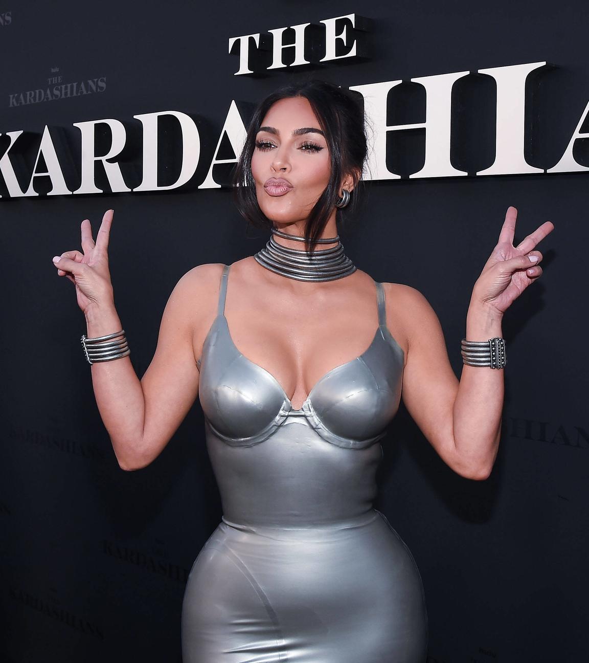 Kim Kardashian steht vor einer schwarzen Wand mit der Aufschrift "The Kardashians". Sie trägt ein silberfarbenes Kleid und silbernen Schmuck. Sie macht einen Kussmund und zeigt mit beiden Händen das Peace-Zeichen.