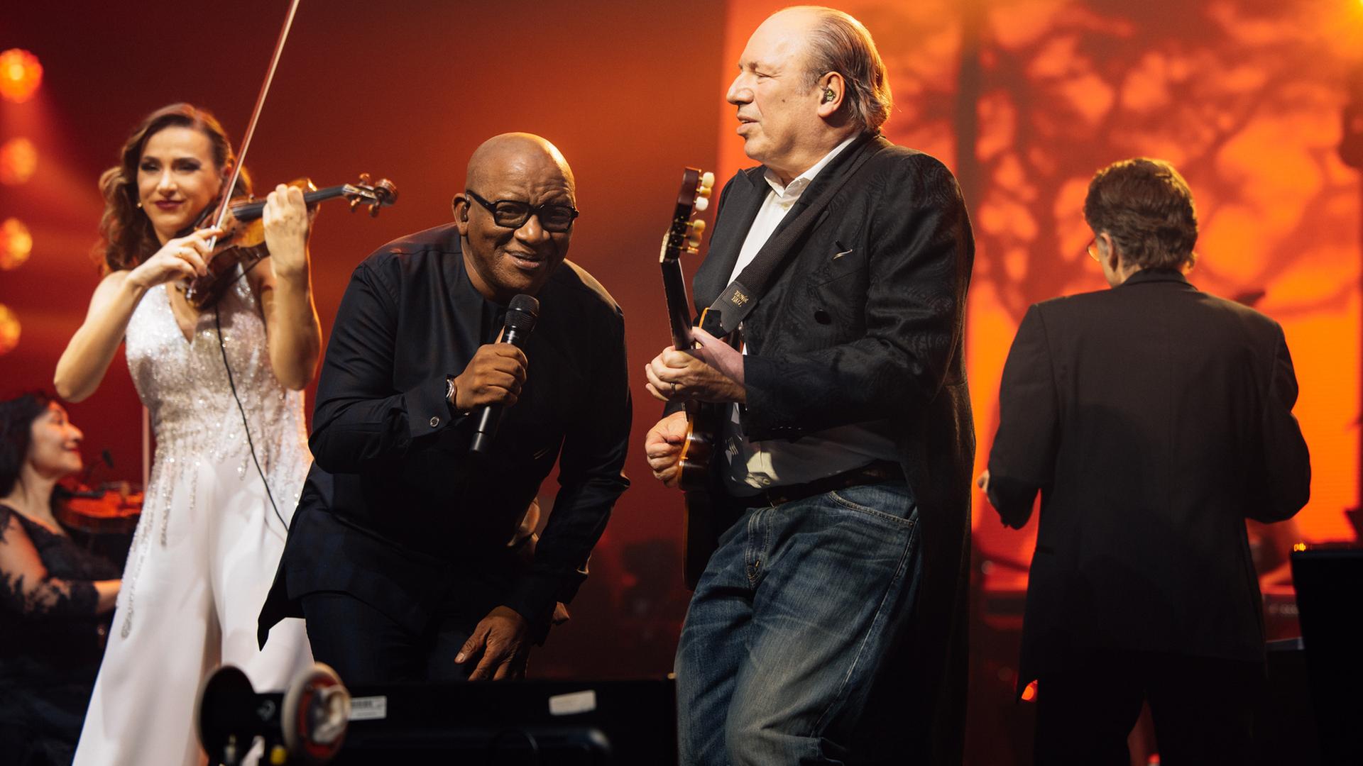 Auf der Buehne: Nebeneinander stehen die Geigerin Rusanda Panfili, der Saenger Lebo M, Hans Zimmer mit Gitarre und, zum Orchester gedreht rueckseitig zu sehen, Dirigent Gavin Greenaway.