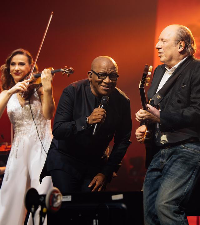 Auf der Buehne: Nebeneinander stehen die Geigerin Rusanda Panfili, der Saenger Lebo M, Hans Zimmer mit Gitarre und, zum Orchester gedreht rueckseitig zu sehen, Dirigent Gavin Greenaway.