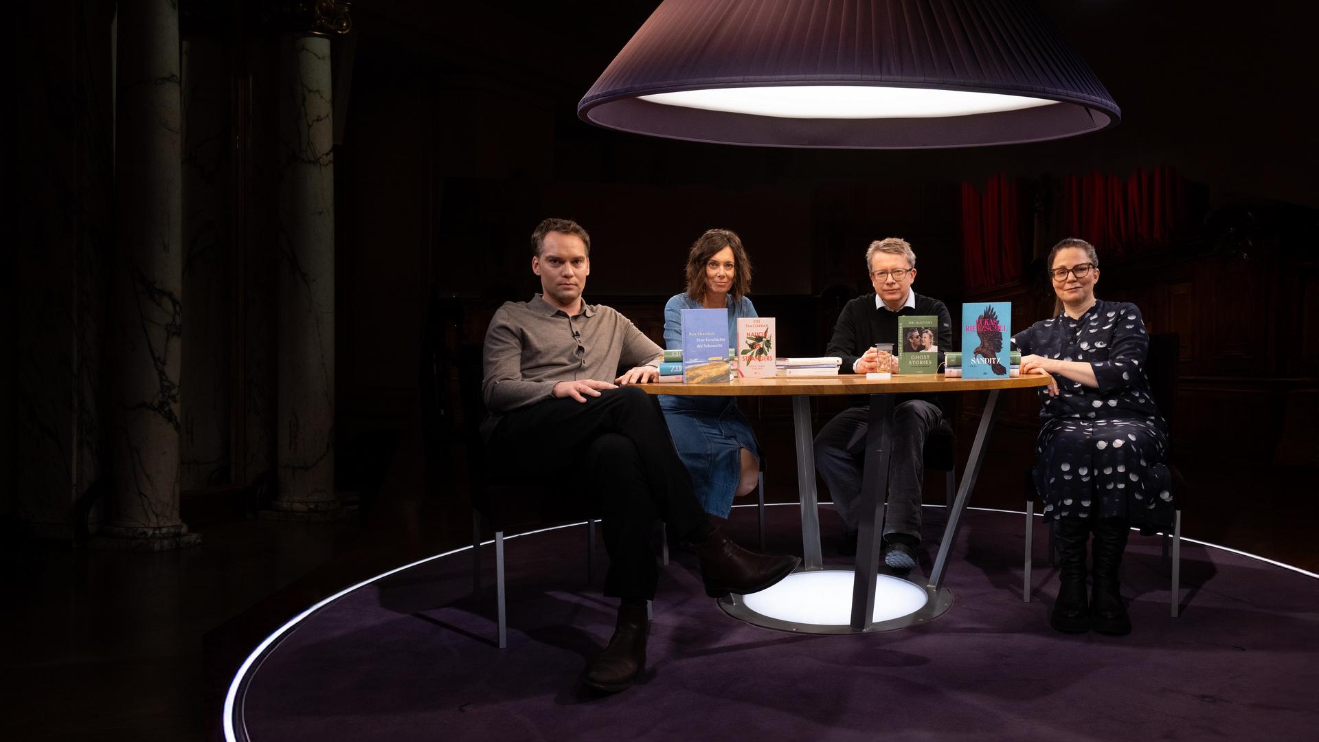 Um einen runden Tisch sitzen zwei Männer und zwei Frauen. Alle blicken in Richtung des Beobachters und vier Bücher stehen auf dem Tisch.