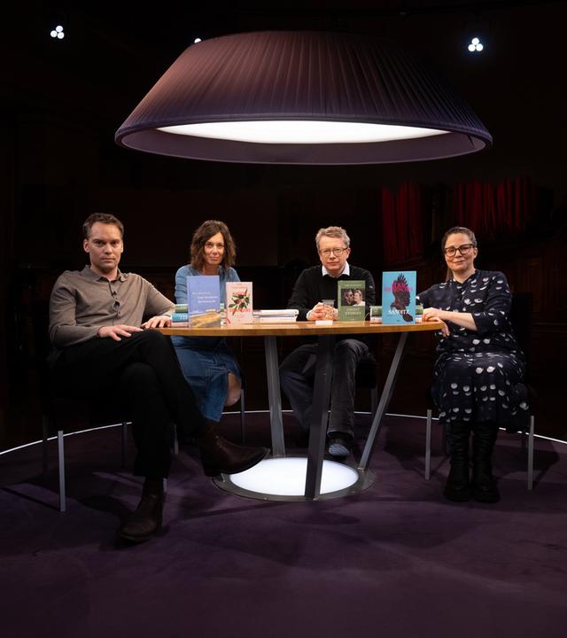 Um einen runden Tisch sitzen zwei Männer und zwei Frauen. Alle blicken in Richtung des Beobachters und vier Bücher stehen auf dem Tisch.