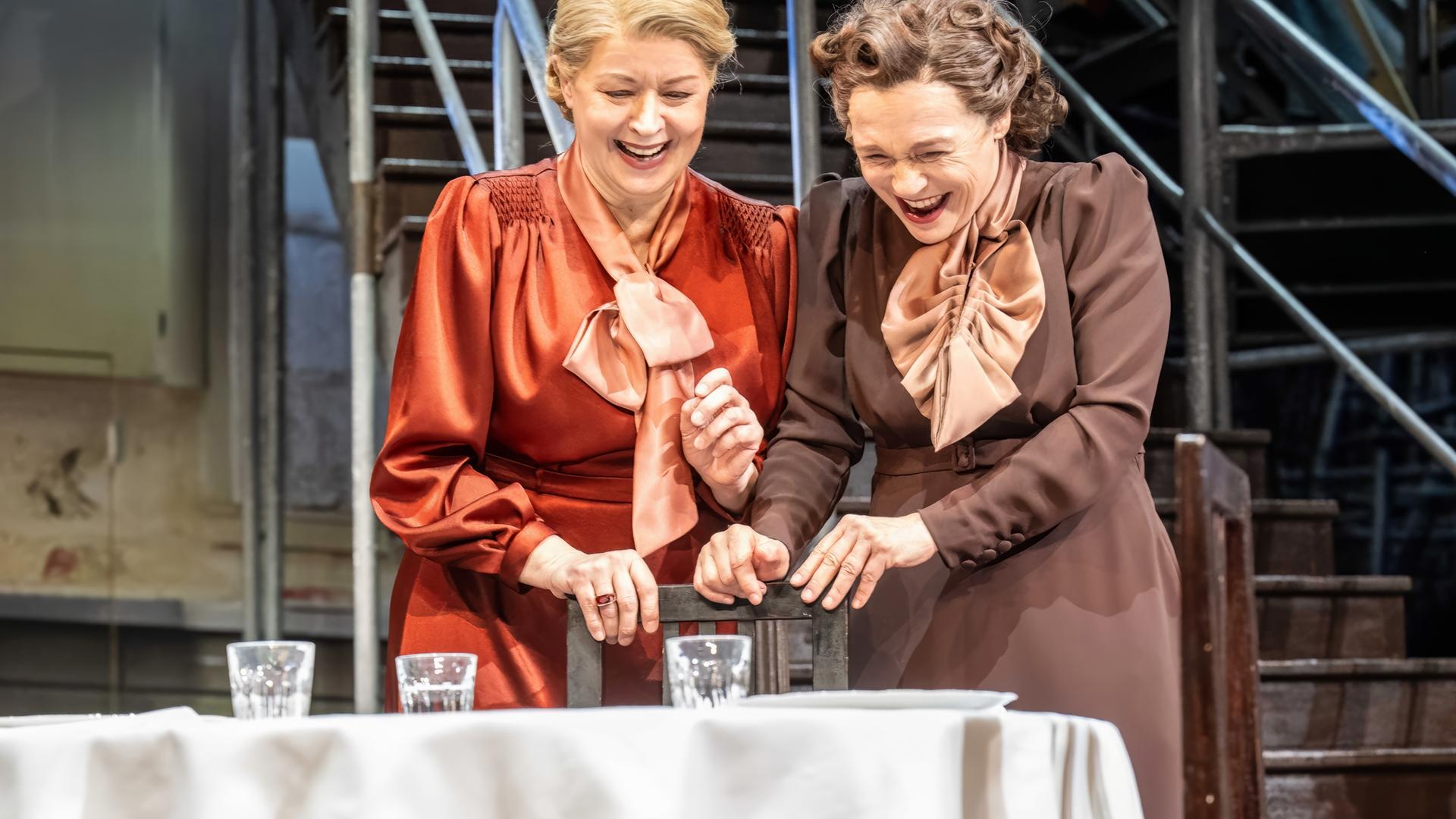 Bühnenszene: Zwei Frauen stehen vor einem gedeckten Tisch und lachen.