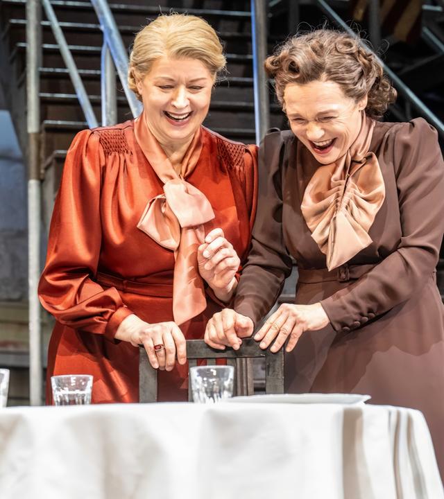 Bühnenszene: Zwei Frauen stehen vor einem gedeckten Tisch und lachen.