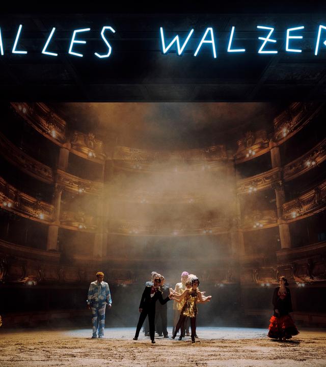 In einer Leuchtschrift und in Großbuchtsaben steht oben im Bild: "Alles Walzer!". Im Hintergrund ist der Zuschauersaal der Wiener Volksoper zu erkennen und im Vordergrund unten ist die Bühne, auf der mehrere Personen in Anzügen und Paillettenkleidern stehen.