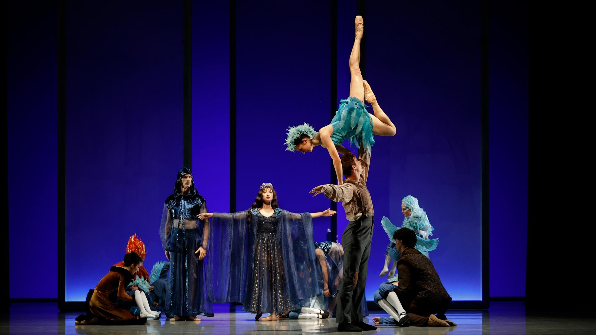 Bühnenszene: Vor blauem Hintergrund performen Tänzerinnen und Tänzer, die schillernde Kostüme tragen, eine Tanzszene.