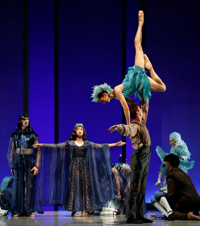 Bühnenszene: Vor blauem Hintergrund performen Tänzerinnen und Tänzer, die schillernde Kostüme tragen, eine Tanzszene.