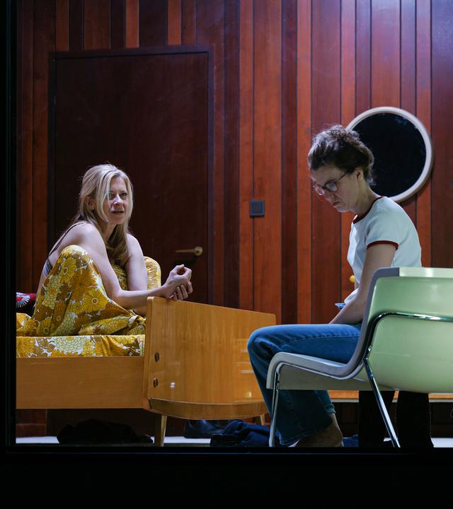 Bühnenszene: Eine Frau sitzt auf einem Bett und spricht mit einer anderen Frau, die auf einem Sessel davor sitzt.