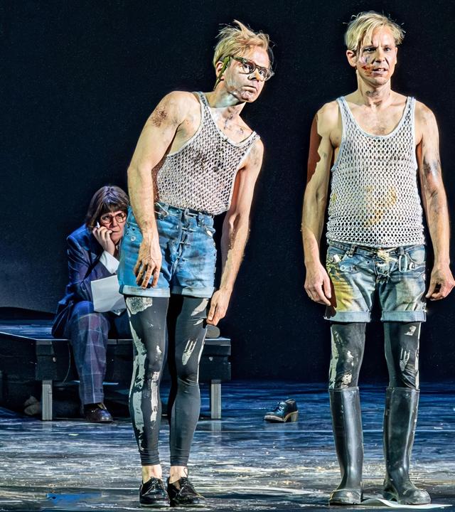 Zwei Männer in kurzen Hosen stehen auf einer Bühne. Im Hintergrund befinden sich zwei weitere Personen und Musikinstrumente.