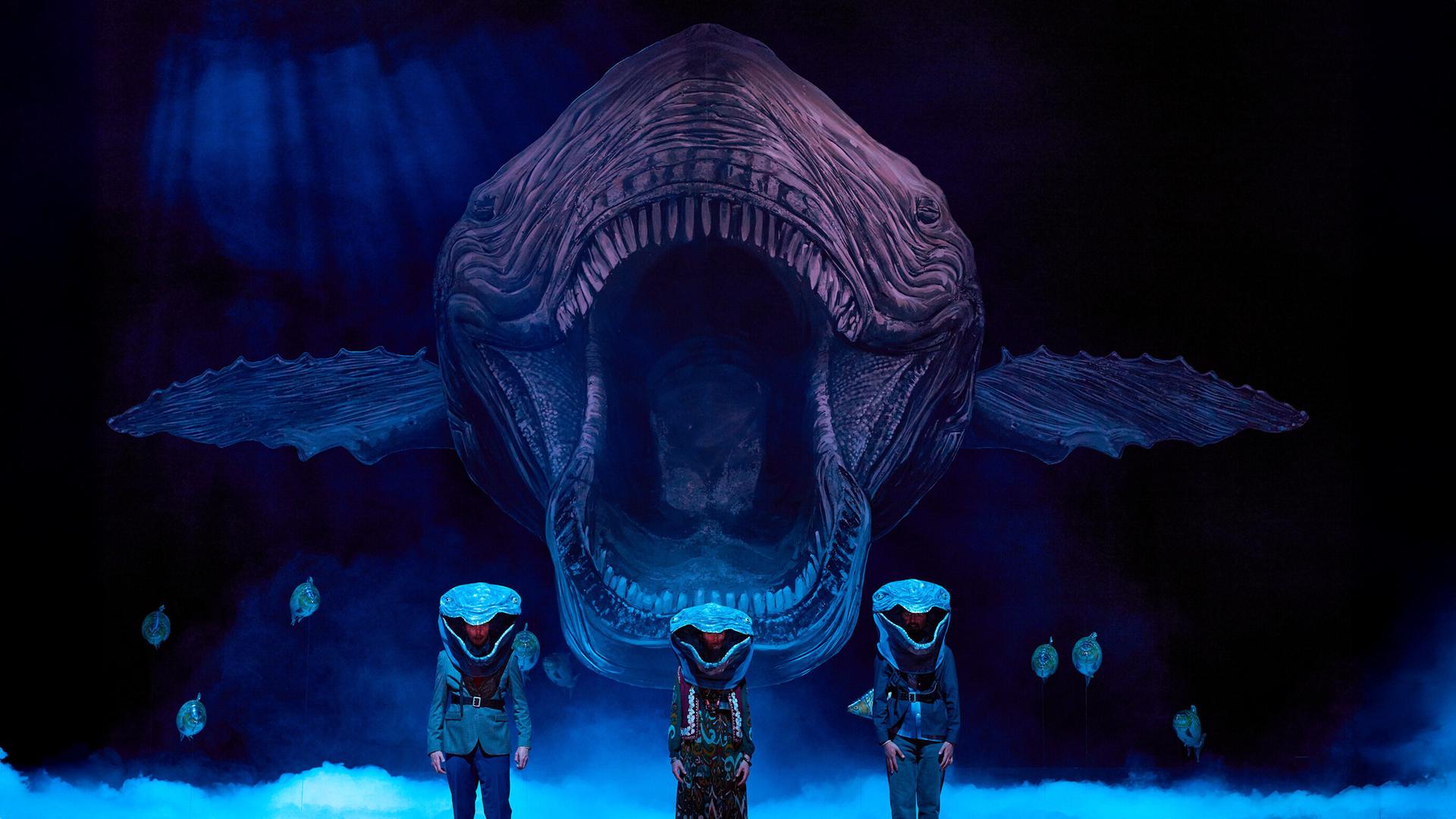 Bühnenszene: Drei Menschen, die als Wale verkleidet sind, stehen vor einem Hintergrund, auf dem ein sehr großer Wal von vorne abgebildet ist.