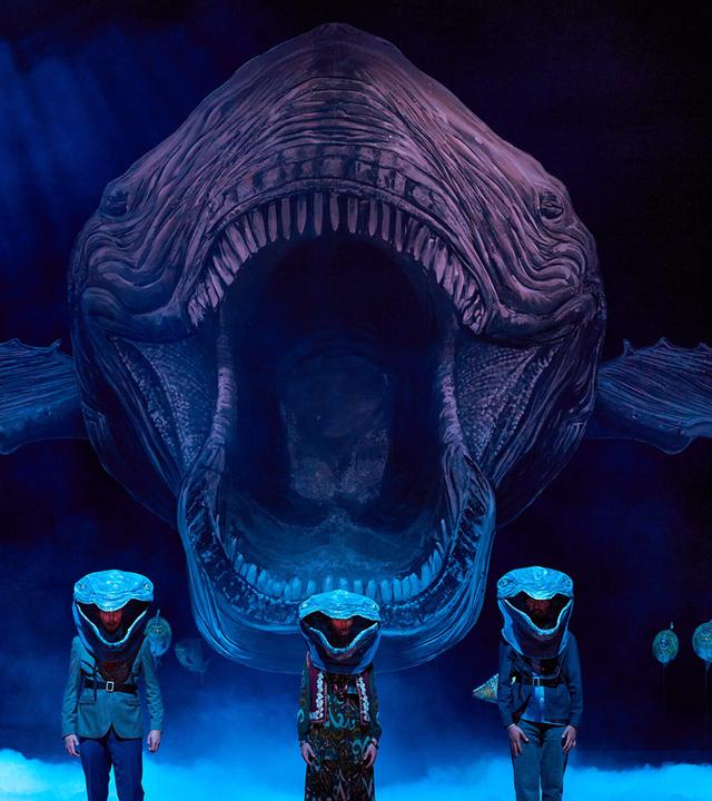 Bühnenszene: Drei Menschen, die als Wale verkleidet sind, stehen vor einem Hintergrund, auf dem ein sehr großer Wal von vorne abgebildet ist.