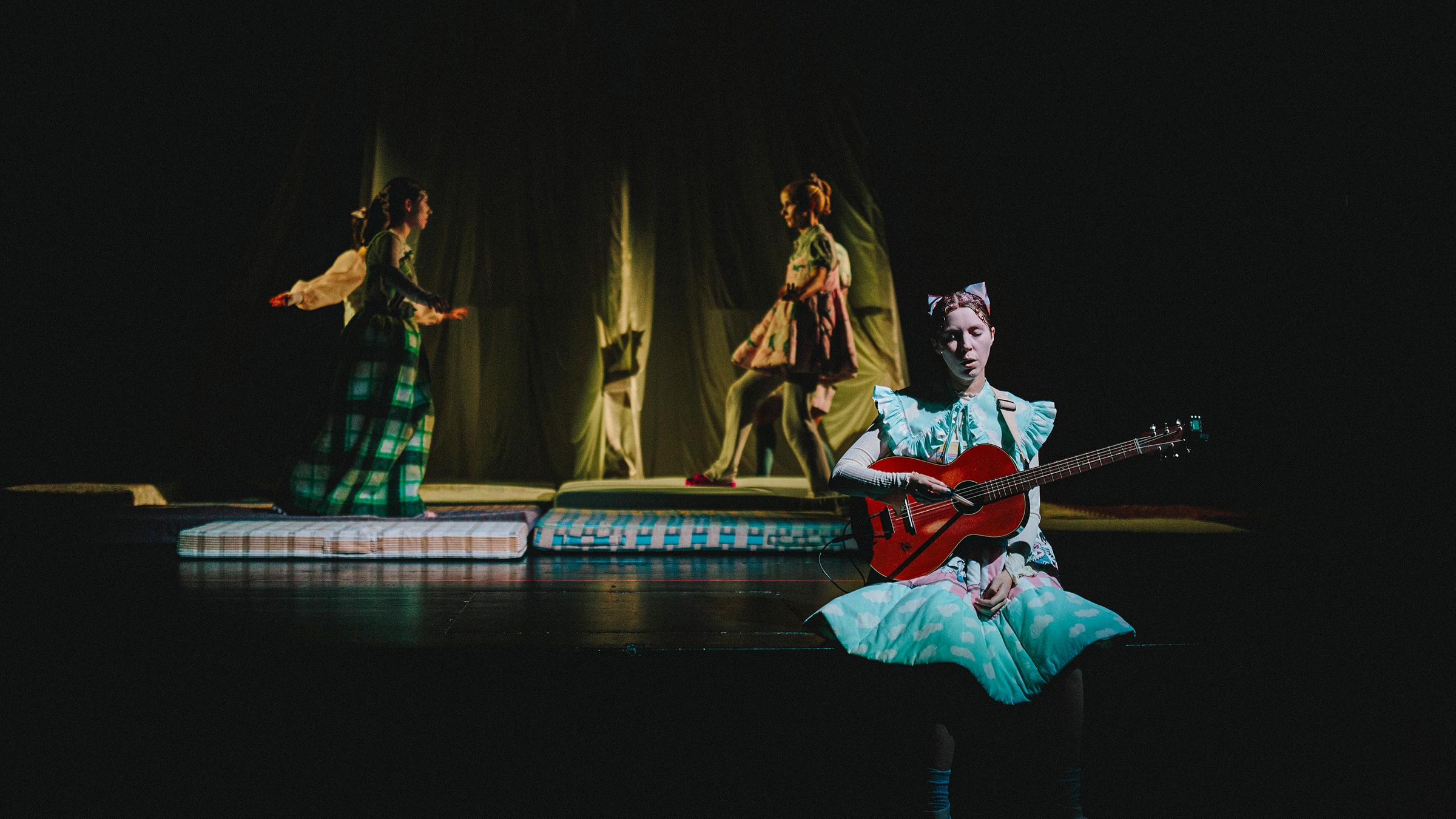 Auf einer dunklen Theaterbühne liegen große Matratzen, darauf stehen sich drei junge Frauen gegenüber. Im Vordergrund sitzt eine Frau im Rüschenkleid mit einer Gitarre und singt oder spricht.