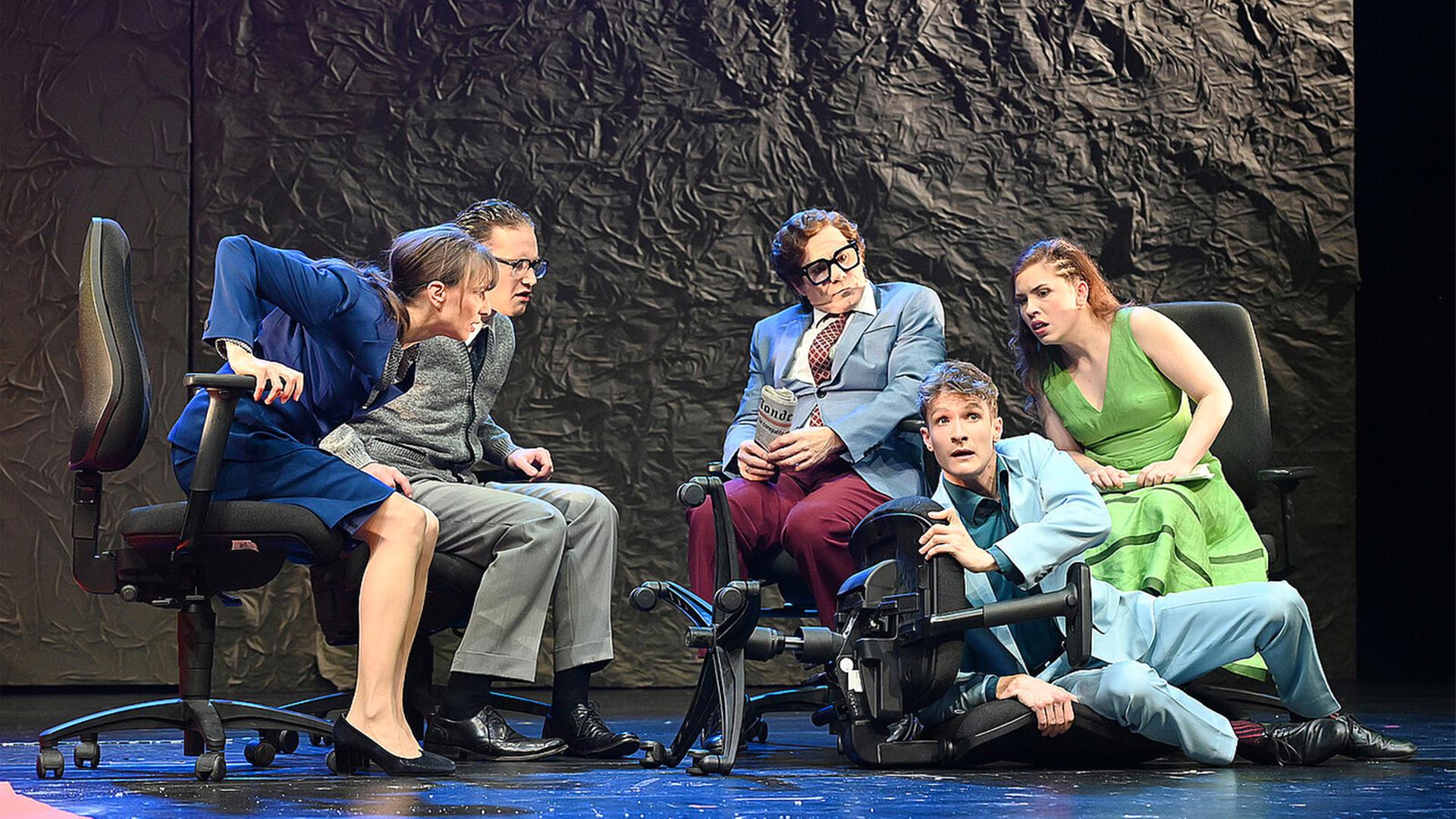 Bühnenszene: Mehrere Personen sitzen auf Bürostühlen im Halbkreis und schauen aufgeregt zu einem Mann, der auf dem Boden neben seinem umgefallenen Stuhl liegt.