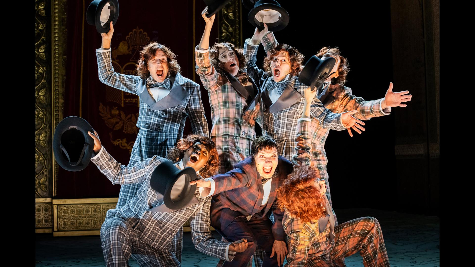 Bühnenszene: Drei Personen knien vor vier stehenden Personen. Alle tragen karierte Anzüge und halten Zylinderhüte in die Luft.