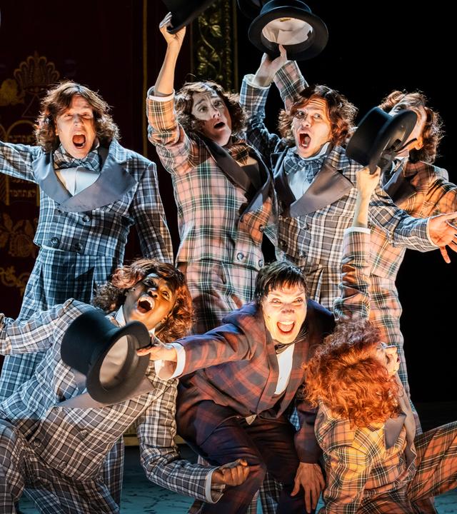 Bühnenszene: Drei Personen knien vor vier stehenden Personen. Alle tragen karierte Anzüge und halten Zylinderhüte in die Luft.