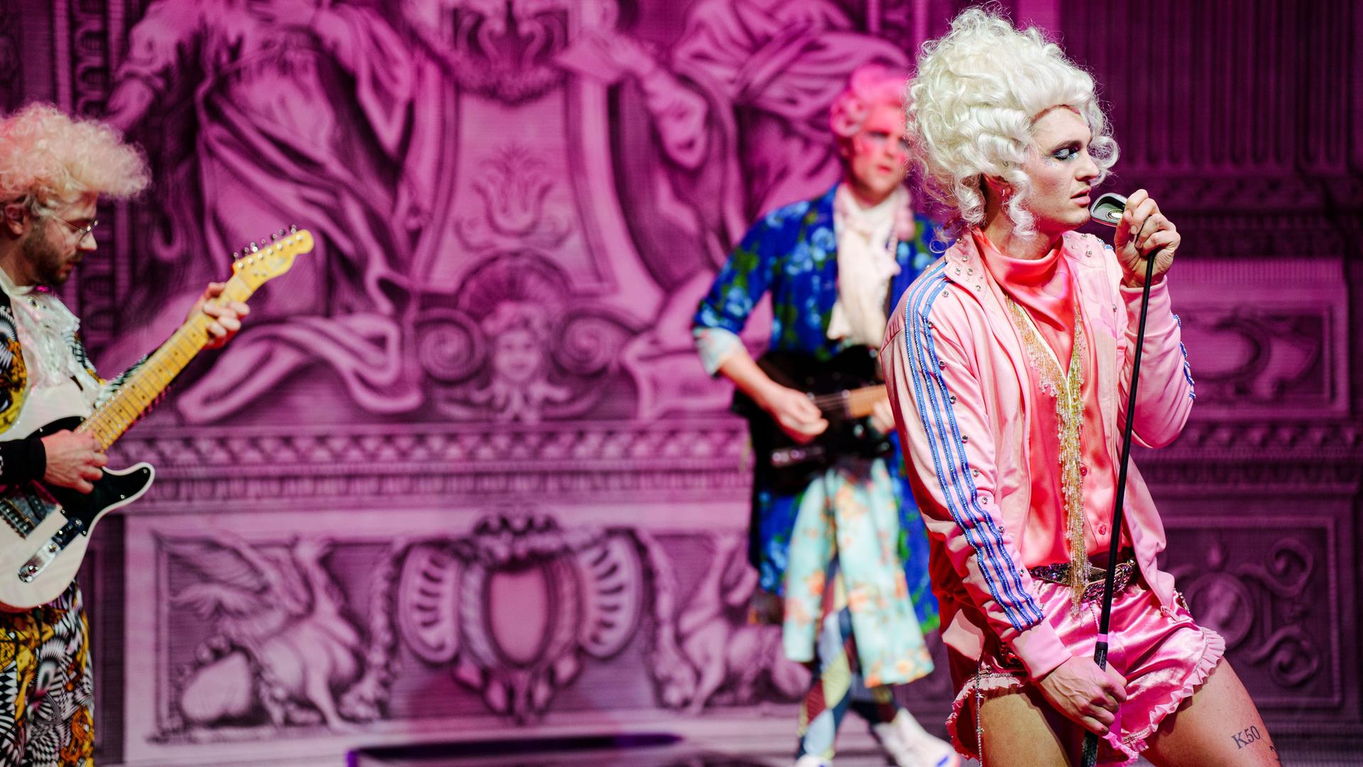 Bühnenszene: Eine Rockband, die Rokoko-Perücken und bunte Sportkleidung trägt spielt vor barocker Kulisse.