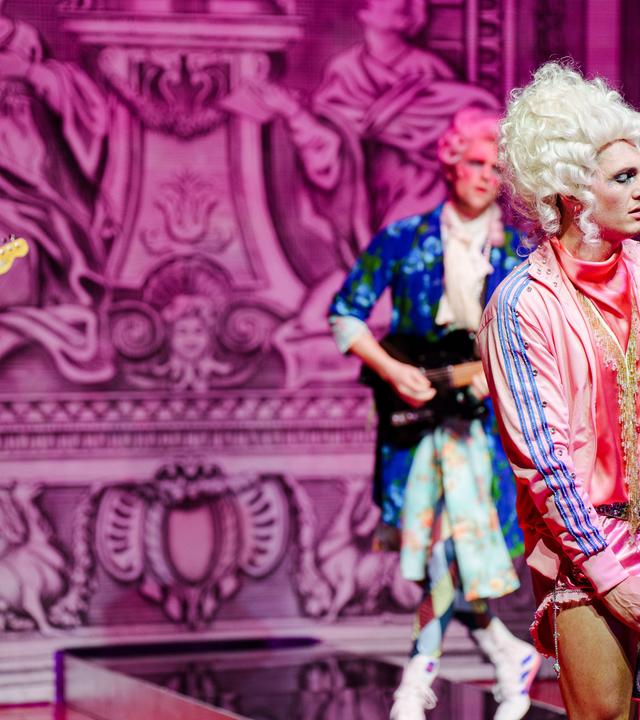 Bühnenszene: Eine Rockband, die Rokoko-Perücken und bunte Sportkleidung trägt spielt vor barocker Kulisse.