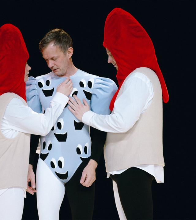 Bühnenszene: Ein Mann in hellblauem Turnanzug ist umgeben von zwei Personen, die rote Kopfbedeckungen tragen.