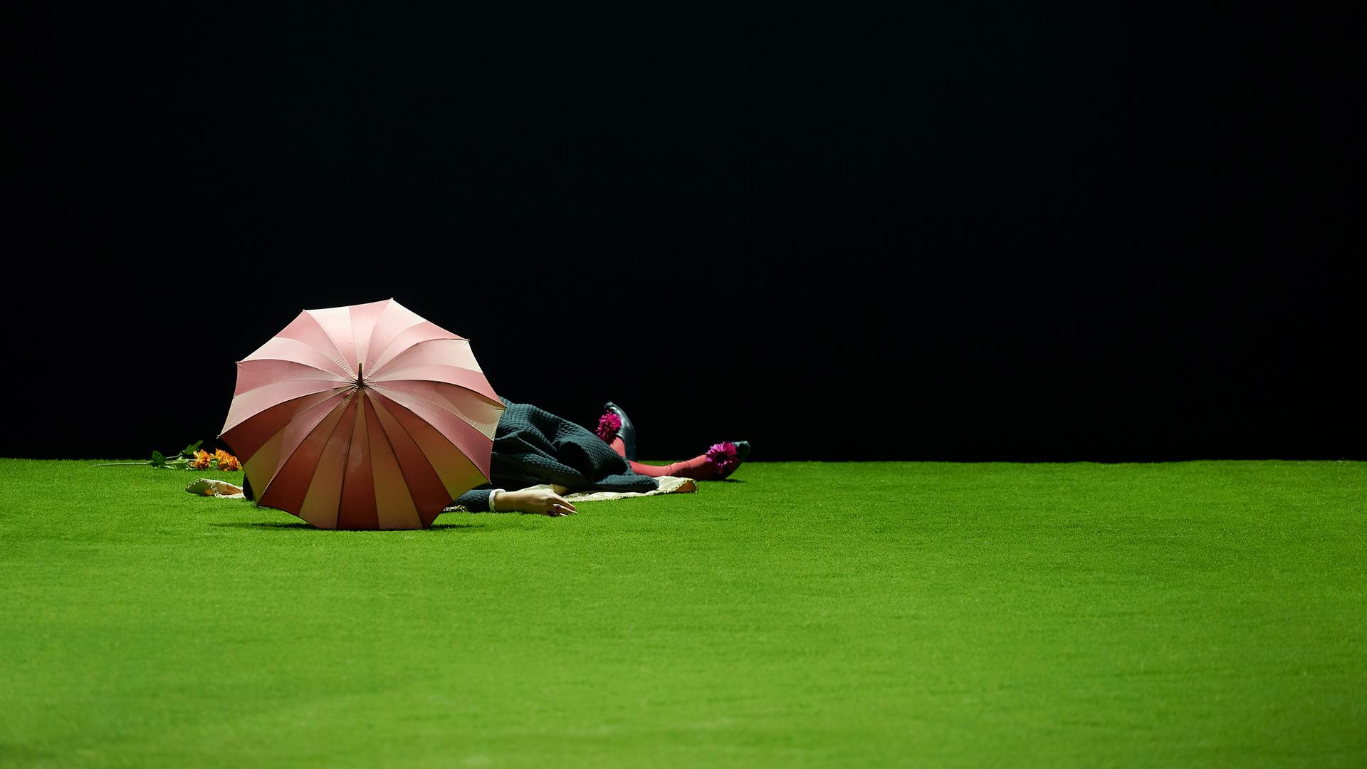 Bühnenszene: Eine Person liegt hinter einem geöffneten Regenschirm auf einem Kunstrasen.