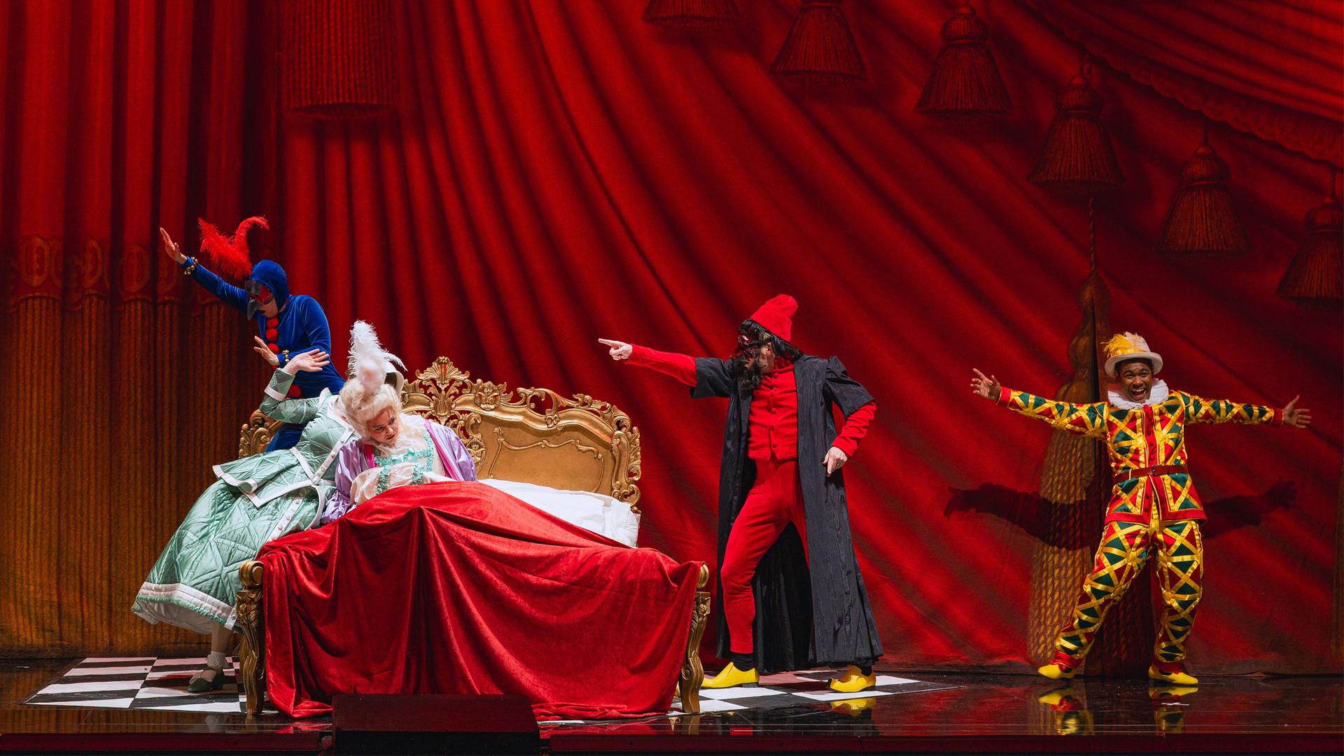 Bühnenszene: Vor einem schweren, roten Vorhang sitzen zwei Frauen in Rokoko-Kleidern auf einem goldenen Bett in barockem Stil. Sie sind umgeben von drei Männern in bunten Kostümen, die jeweils mit dem rechten Arm nach links zeigen.