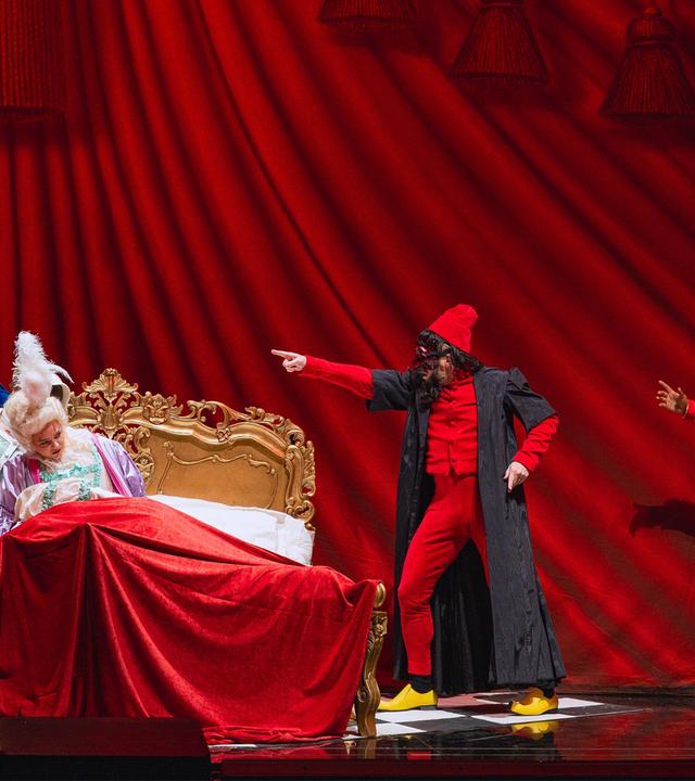 Bühnenszene: Vor einem schweren, roten Vorhang sitzen zwei Frauen in Rokoko-Kleidern auf einem goldenen Bett in barockem Stil. Sie sind umgeben von drei Männern in bunten Kostümen, die jeweils mit dem rechten Arm nach links zeigen.