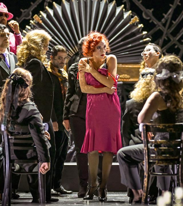 Bühnenszene: Eine Frau in einem pinkfarbenen Kleid ist umringt von Männern und Frauen.