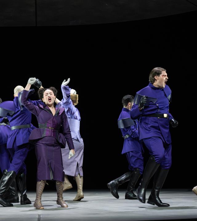 Bühnenszene: Menschen in violett-farbener Bekleidung tanzen auf einer Bühne.