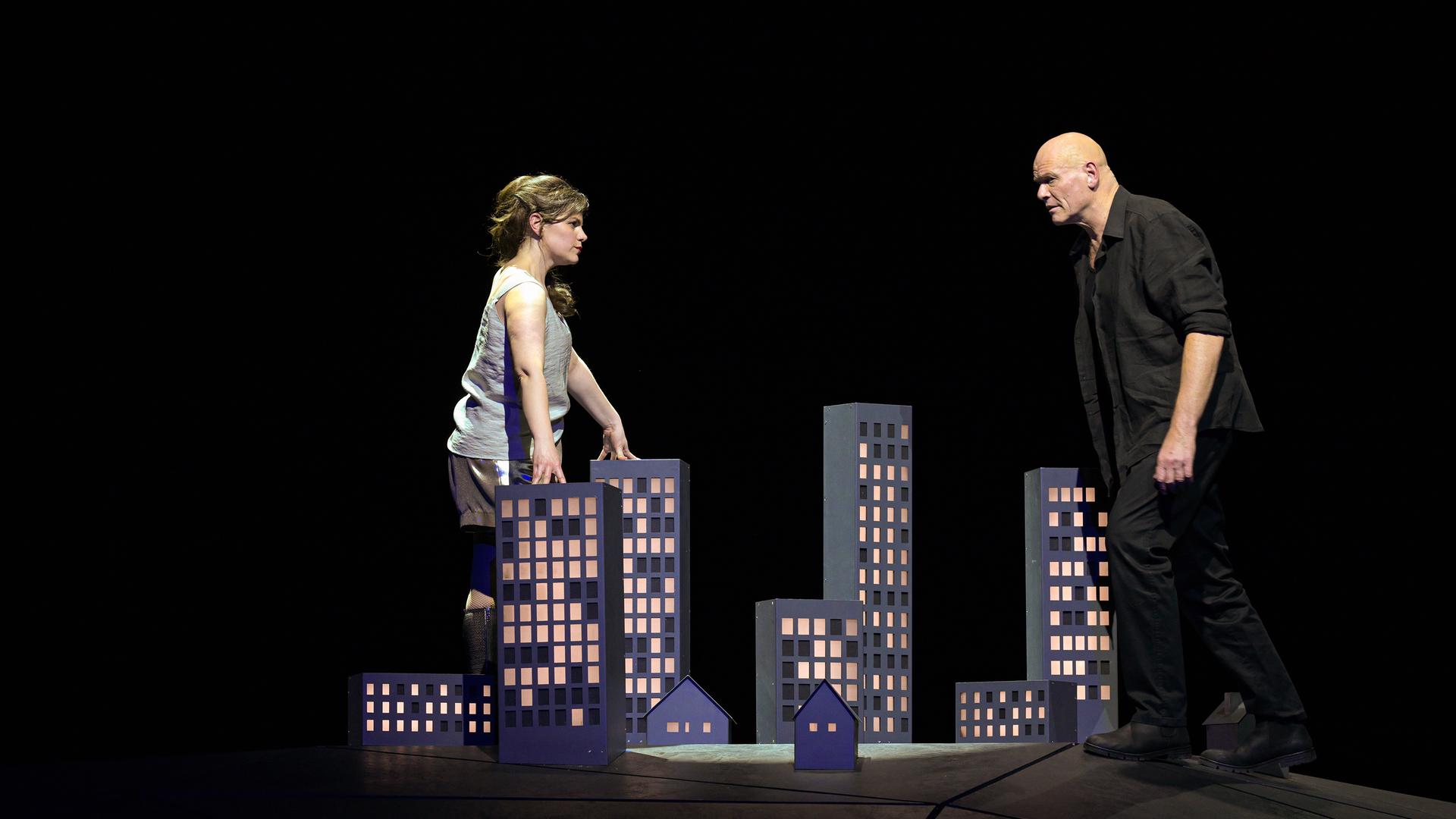 Bühnenszene: Eine Frau und ein Mann stehen sich gegenüber und sehen sich an. Zwischen ihnen steht auf dem Boden das Modell einer Stadt.