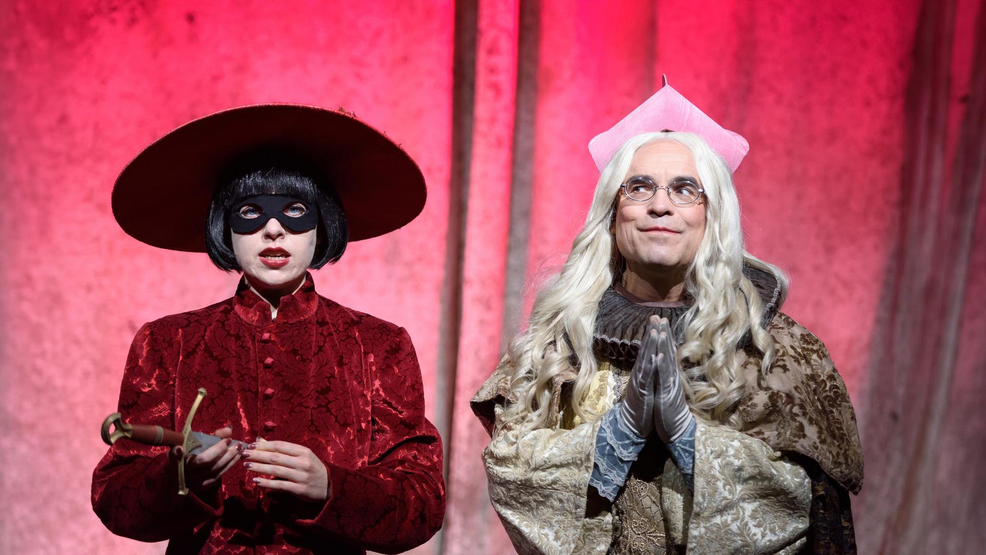 Bühnenszene: Eine Frau mit Maske und großem Hut steht links neben einem Mann in geistlichem Kostüm, vor pinkfarbenem Hintergrund.