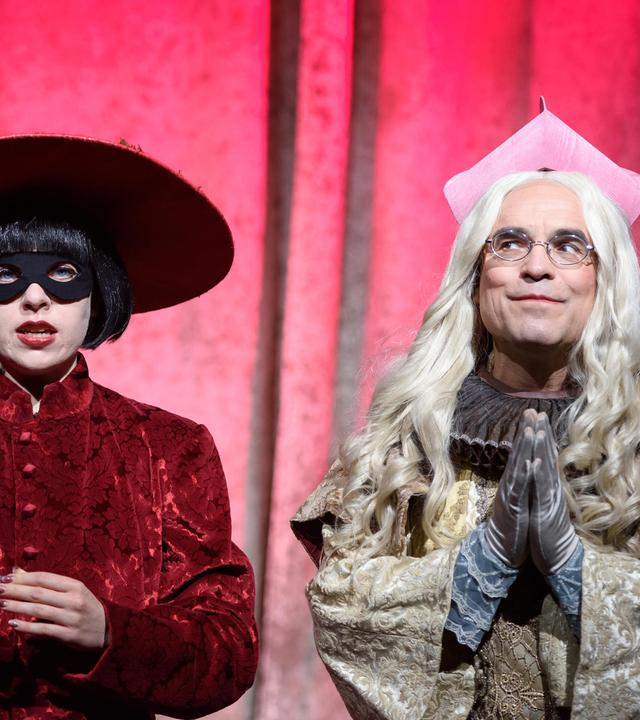 Bühnenszene: Eine Frau mit Maske und großem Hut steht links neben einem Mann in geistlichem Kostüm, vor pinkfarbenem Hintergrund.
