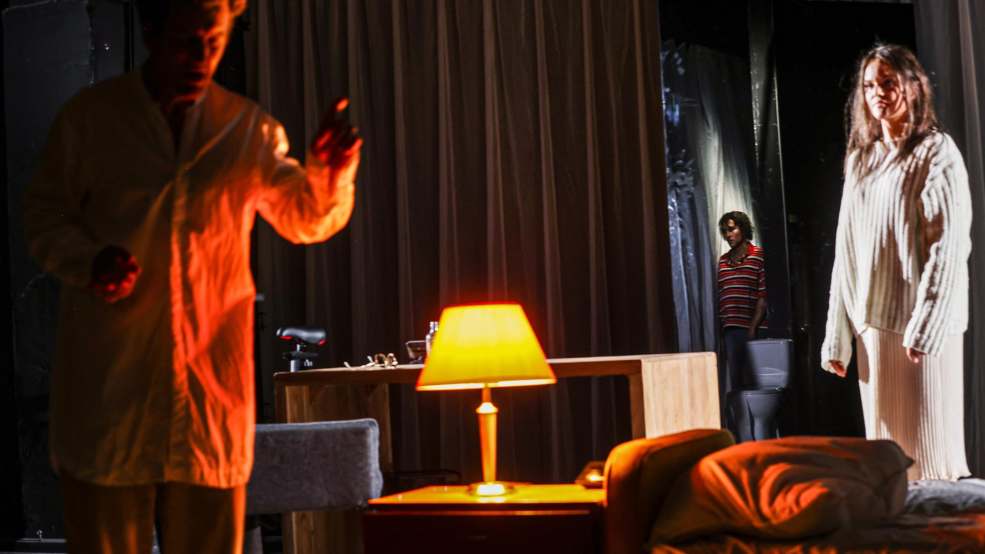 Bühnenszene: In einem abgedunkelten Wohnzimmer diskutieren ein Mann und eine Frau. Ein Junge beobachtet die Szene aus der Entfernung.