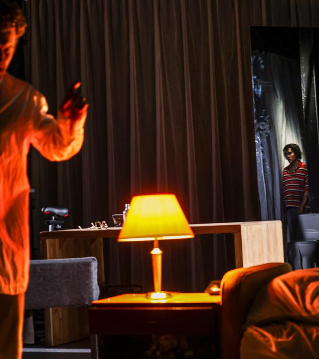 Bühnenszene: In einem abgedunkelten Wohnzimmer diskutieren ein Mann und eine Frau. Ein Junge beobachtet die Szene aus der Entfernung.