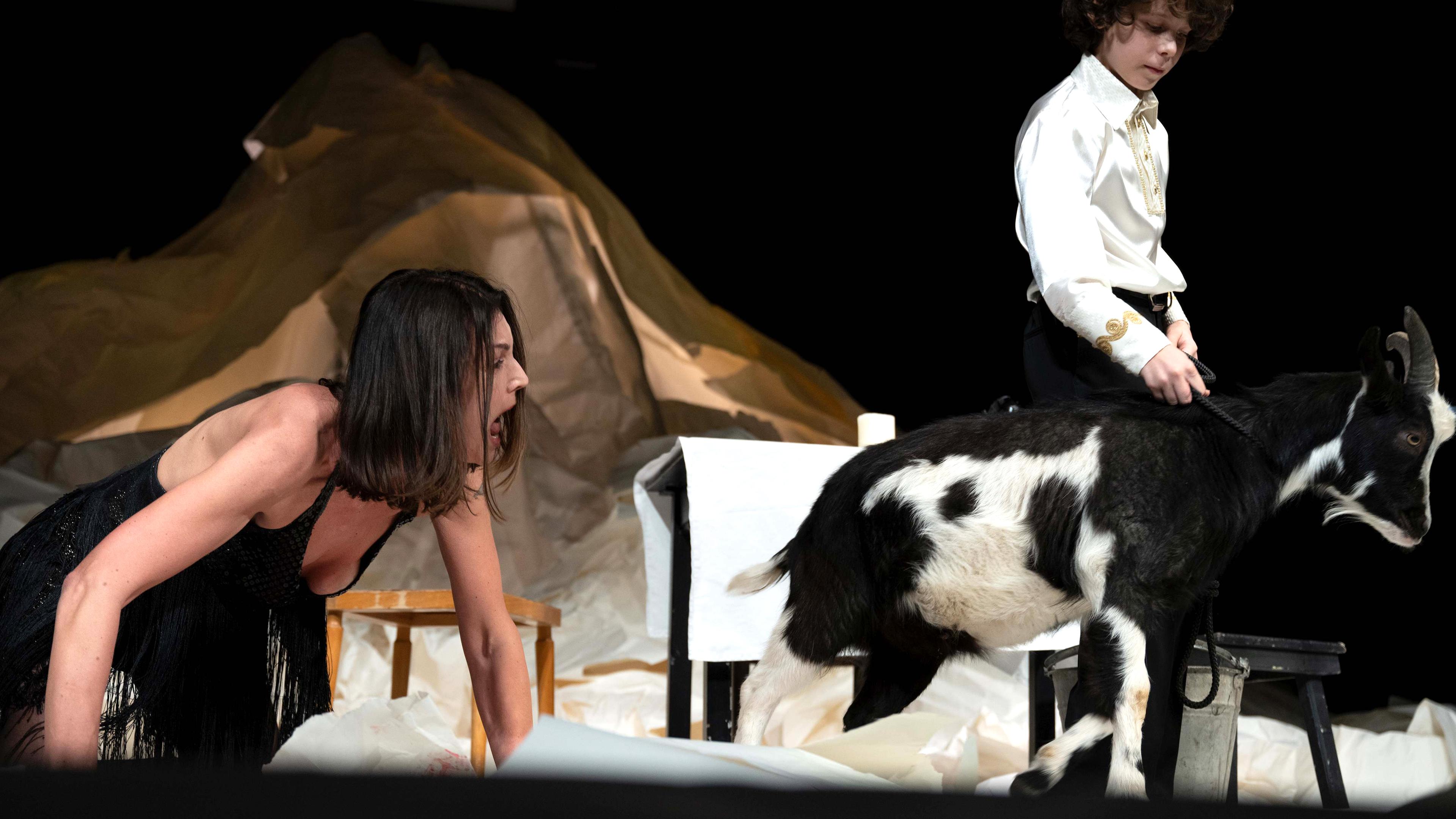 Bühnenszene: Eine Frau kauert auf dem Boden und schreit eine Ziege an, die ein Junge an der Leine führt.