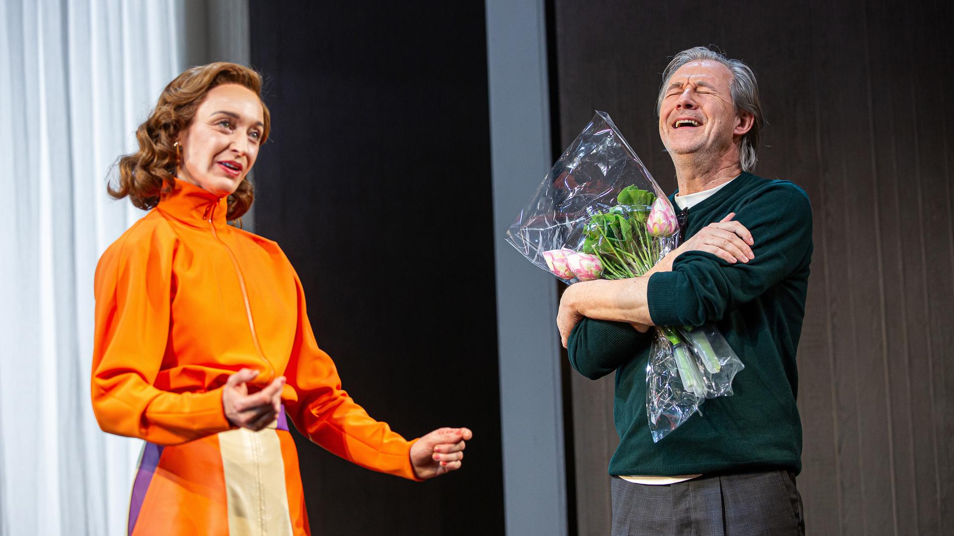 Bühnenszene: Eine Frau in orange-farbenem Kleid steht neben einem Mann, der einen Blumenstrauß in den Armen hält.