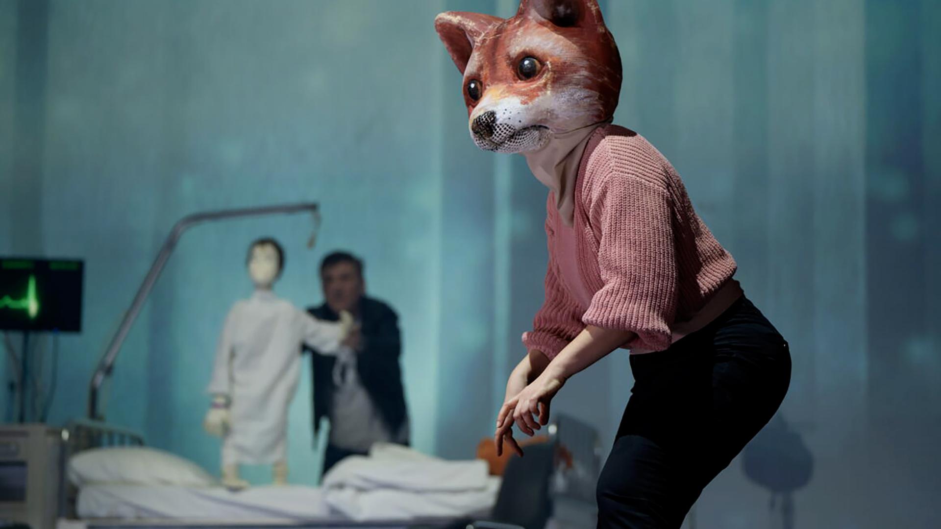 Bühnenszene: Ein Mensch, der eine große Fuchsmaske trägt, steht vor einem Krankenbett. Hinter dem Krankenbett steht ein Mann, der eine Puppe in Form eines Kindes hält.