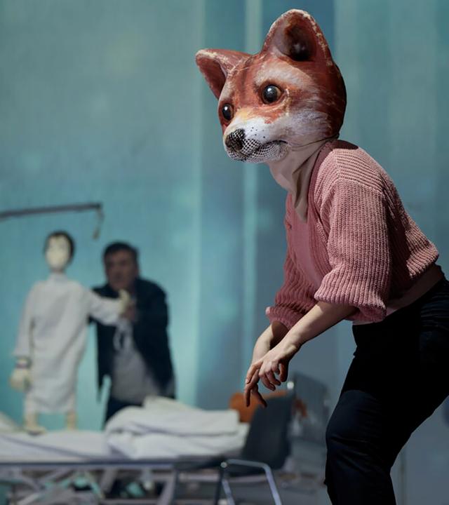 Bühnenszene: Ein Mensch, der eine große Fuchsmaske trägt, steht vor einem Krankenbett. Hinter dem Krankenbett steht ein Mann, der eine Puppe in Form eines Kindes hält.