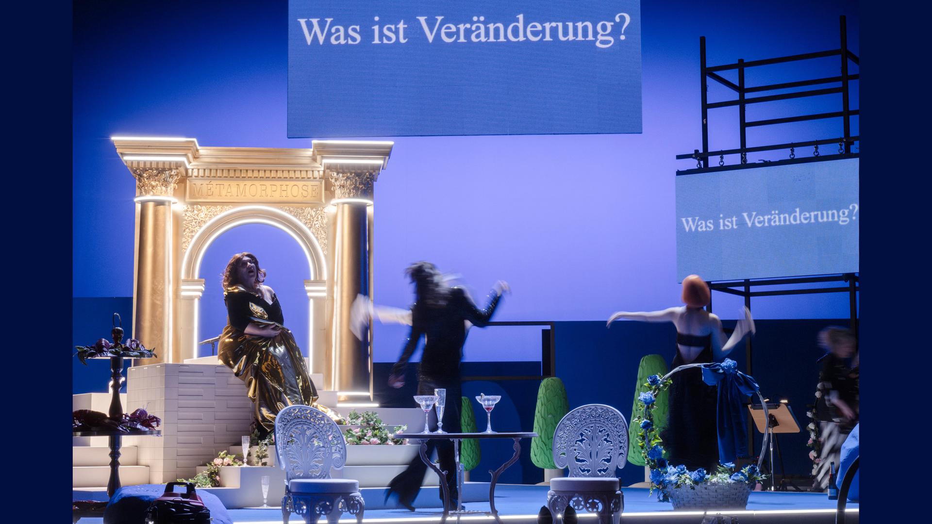 Bühnenszene: Drei Frauen sitzen und tanzen in einem Wohnzimmer. Im Hintergrund ist ein Portal zu sehen. Oben hängt ein Schild mit einer Aufschrift. Die Bühne ist blau angeleuchtet.