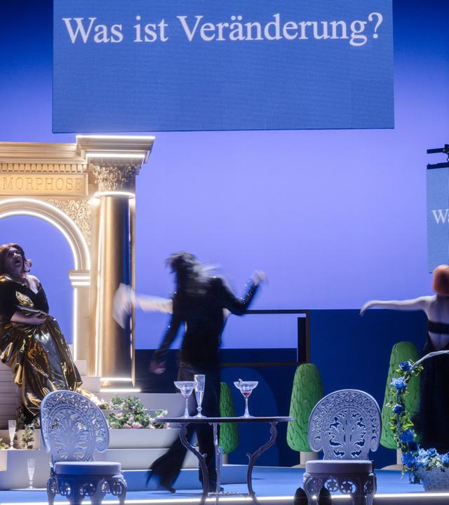 Bühnenszene: Drei Frauen sitzen und tanzen in einem Wohnzimmer. Im Hintergrund ist ein Portal zu sehen. Oben hängt ein Schild mit einer Aufschrift. Die Bühne ist blau angeleuchtet.