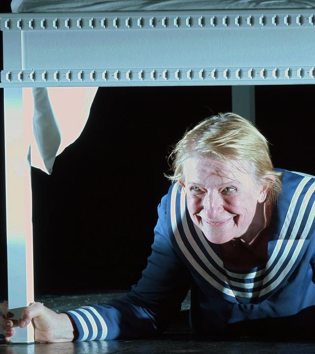 „THEATER.MACHER.INNEN“: Dagmar Manzel liegt in einem Matrosen-Kostüm und einem weißen Tisch und hält mit beiden Händen zwei Tischbeine in der Hand. Sie grinst verschmitzt nach vorne.