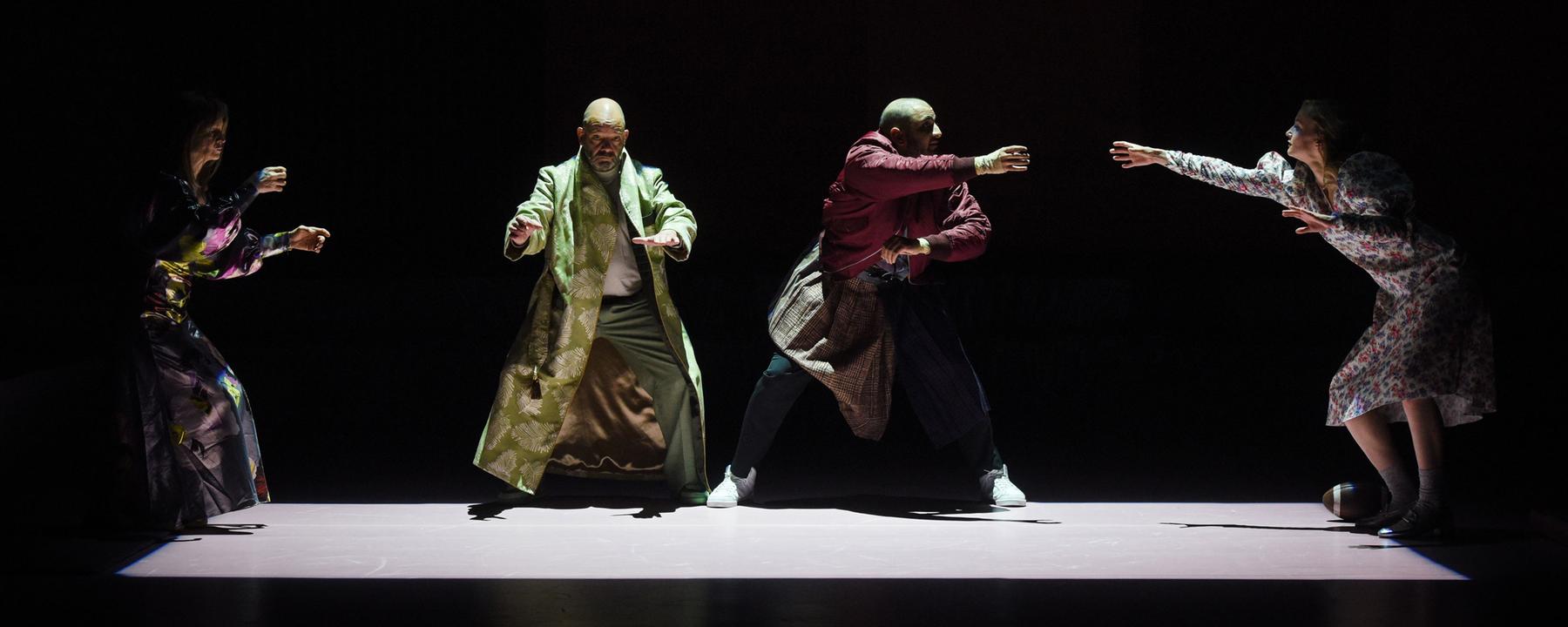 : Auf einer dunklen Bühne ist mit Licht auf dem Boden ein Rechteck abgegrenzt. Links an der schmalen Seite steht eine Frau mit auf Höhe der Hüfte erhobenen Armen, sie trägt ein glänzendes, buntgemustertes langes Kleid. Rechts neben ihr, nach vorne gewandt, ebenfalls mit erhobenen Armen steht mit leicht gespreizten Beinen ein glatzköpfiger Mann, er trägt einen langen hellgrünen Mantel. Neben ihm in ähnlicher Position ein weiterer, etwas jüngerer glatzköpfiger Mann, in einer dunkelroten Bomberjacke, einem Schottenrock über der Jeans und weißen Turnschuhen. Er wendet sich nach rechts, dort steht an der anderen Schmalseite eine junge blonde Frau in einem weißen Kleid, mit Blümchenmuster. Sie streckt den rechten Arm zu dem Mann neben ihr aus. Zu ihren Füßen liegt rechts ein Football. 