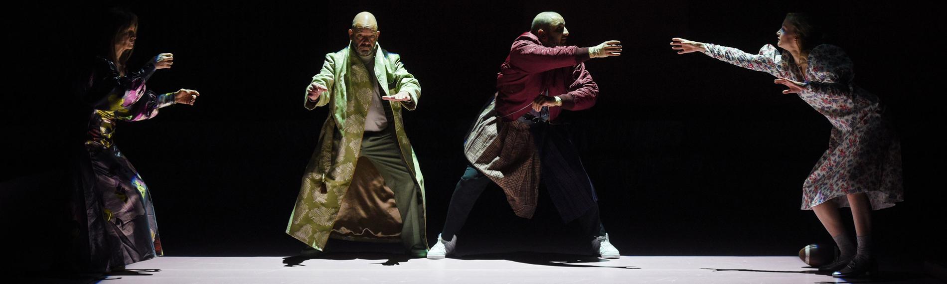 : Auf einer dunklen Bühne ist mit Licht auf dem Boden ein Rechteck abgegrenzt. Links an der schmalen Seite steht eine Frau mit auf Höhe der Hüfte erhobenen Armen, sie trägt ein glänzendes, buntgemustertes langes Kleid. Rechts neben ihr, nach vorne gewandt, ebenfalls mit erhobenen Armen steht mit leicht gespreizten Beinen ein glatzköpfiger Mann, er trägt einen langen hellgrünen Mantel. Neben ihm in ähnlicher Position ein weiterer, etwas jüngerer glatzköpfiger Mann, in einer dunkelroten Bomberjacke, einem Schottenrock über der Jeans und weißen Turnschuhen. Er wendet sich nach rechts, dort steht an der anderen Schmalseite eine junge blonde Frau in einem weißen Kleid, mit Blümchenmuster. Sie streckt den rechten Arm zu dem Mann neben ihr aus. Zu ihren Füßen liegt rechts ein Football. 