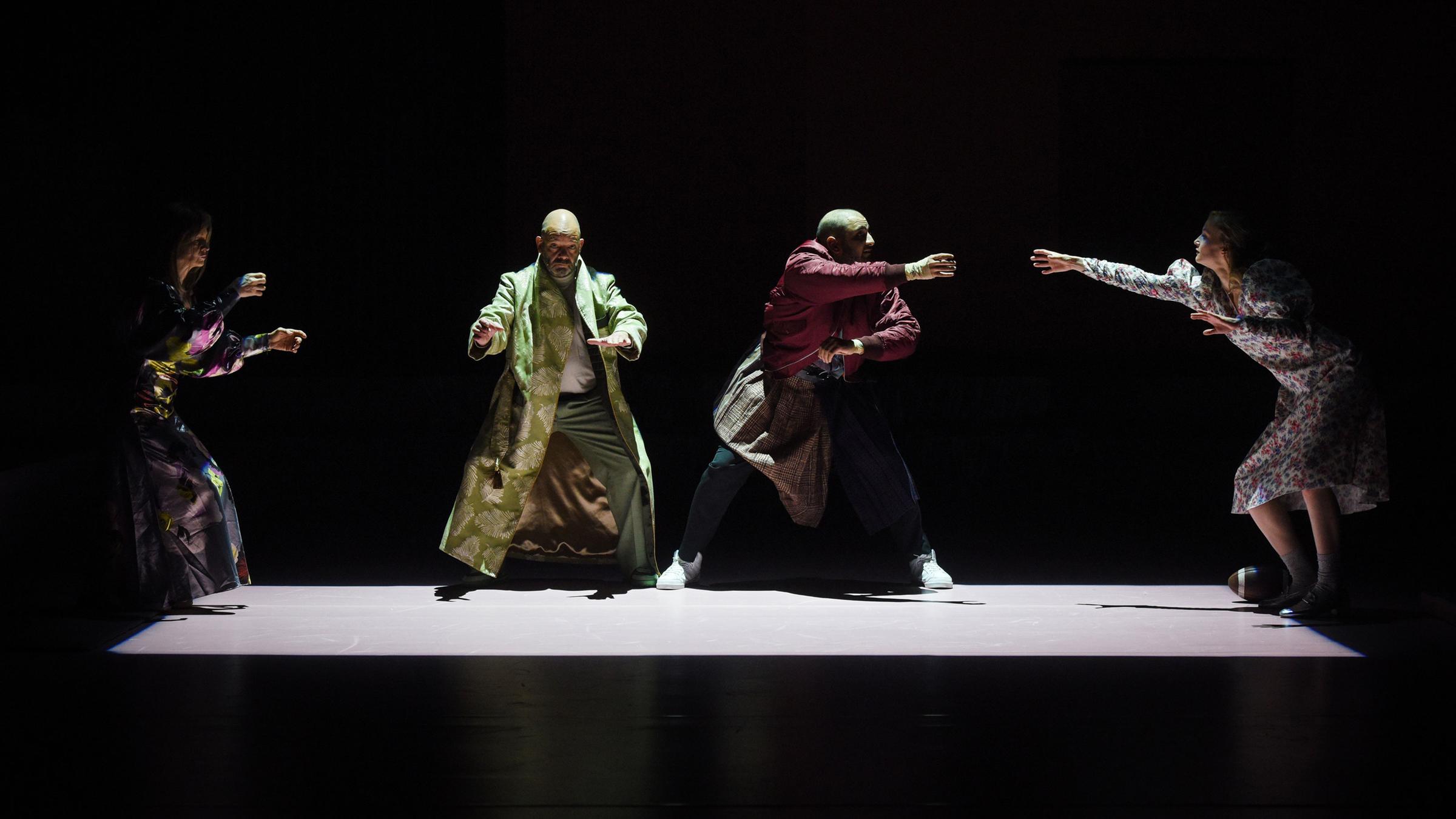 : Auf einer dunklen Bühne ist mit Licht auf dem Boden ein Rechteck abgegrenzt. Links an der schmalen Seite steht eine Frau mit auf Höhe der Hüfte erhobenen Armen, sie trägt ein glänzendes, buntgemustertes langes Kleid. Rechts neben ihr, nach vorne gewandt, ebenfalls mit erhobenen Armen steht mit leicht gespreizten Beinen ein glatzköpfiger Mann, er trägt einen langen hellgrünen Mantel. Neben ihm in ähnlicher Position ein weiterer, etwas jüngerer glatzköpfiger Mann, in einer dunkelroten Bomberjacke, einem Schottenrock über der Jeans und weißen Turnschuhen. Er wendet sich nach rechts, dort steht an der anderen Schmalseite eine junge blonde Frau in einem weißen Kleid, mit Blümchenmuster. Sie streckt den rechten Arm zu dem Mann neben ihr aus. Zu ihren Füßen liegt rechts ein Football. 