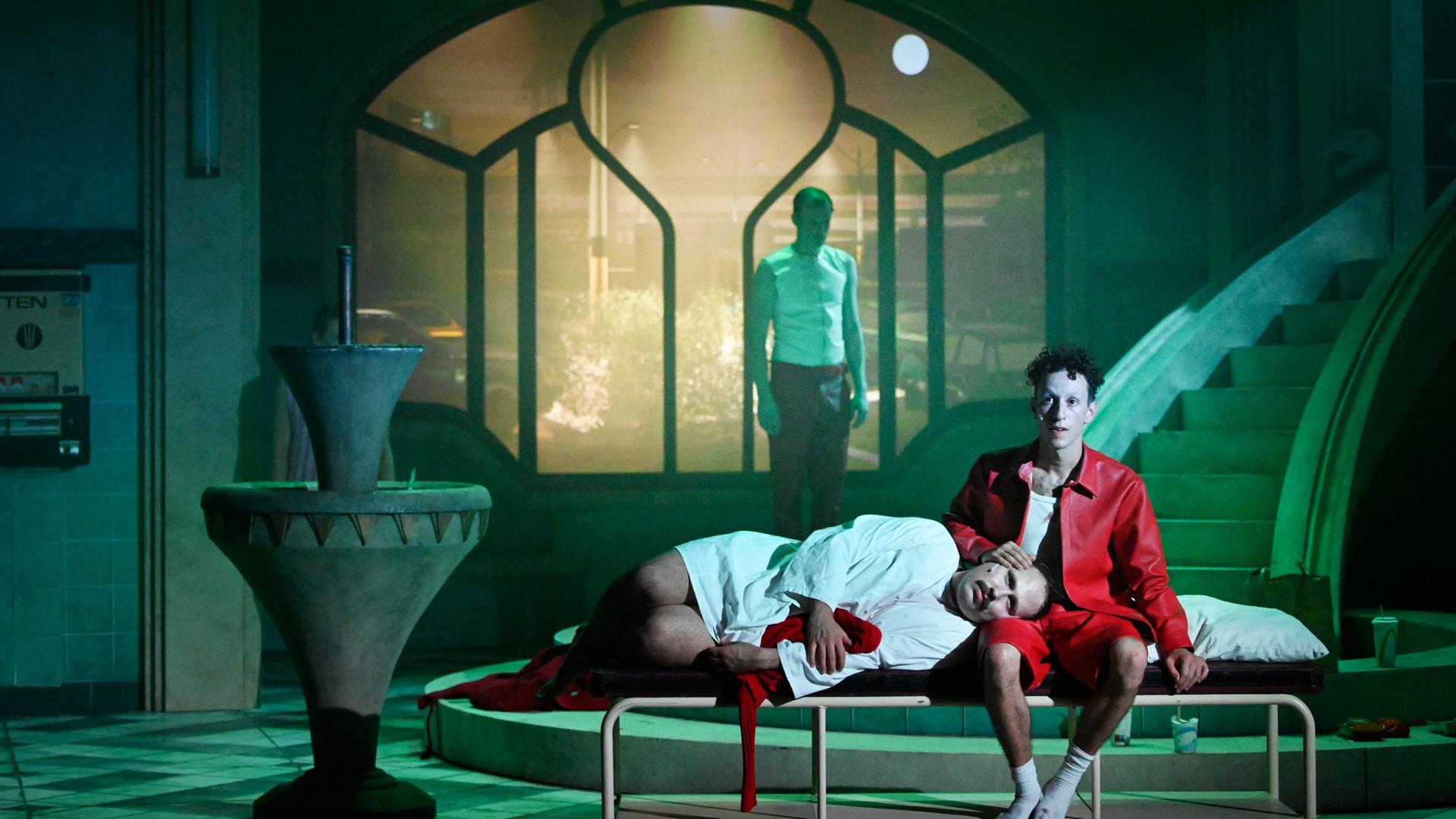 Düstere Theaterszene mit expressionistischem Bühnenbild: Eine Person in einem roten Hemd und roter Hose sitzt barfuß auf einem schlichten Metallbett, mit leerem Blick nach vorne. Eine andere Person in weißem Hemd und dunkler Hose liegt mit dem Kopf auf ihrem Schoß, die Augen geschlossen. Im Hintergrund steht eine weitere Person mit weißem Unterhemd und roter Hose vor einem großen, schlüsselförmigen Fenster, durch das eine nächtliche Stadtszene mit Autos und Straßenlaternen zu sehen ist. Links im Bild ein Brunnen, der Boden ist gefliest. 